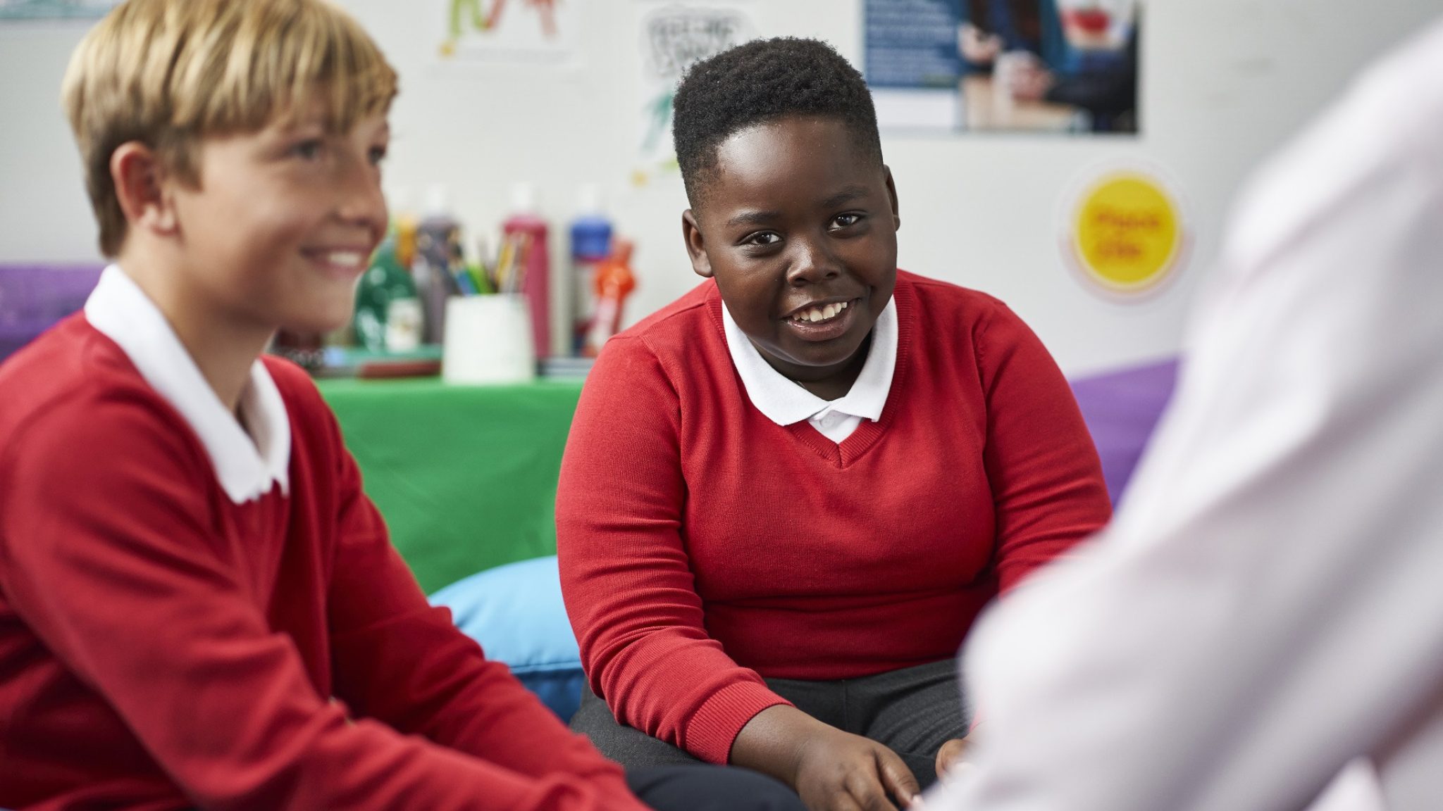 Two boys wearing red jumpers and smiling sit in a classroom talking to an adult whose back is to the camera